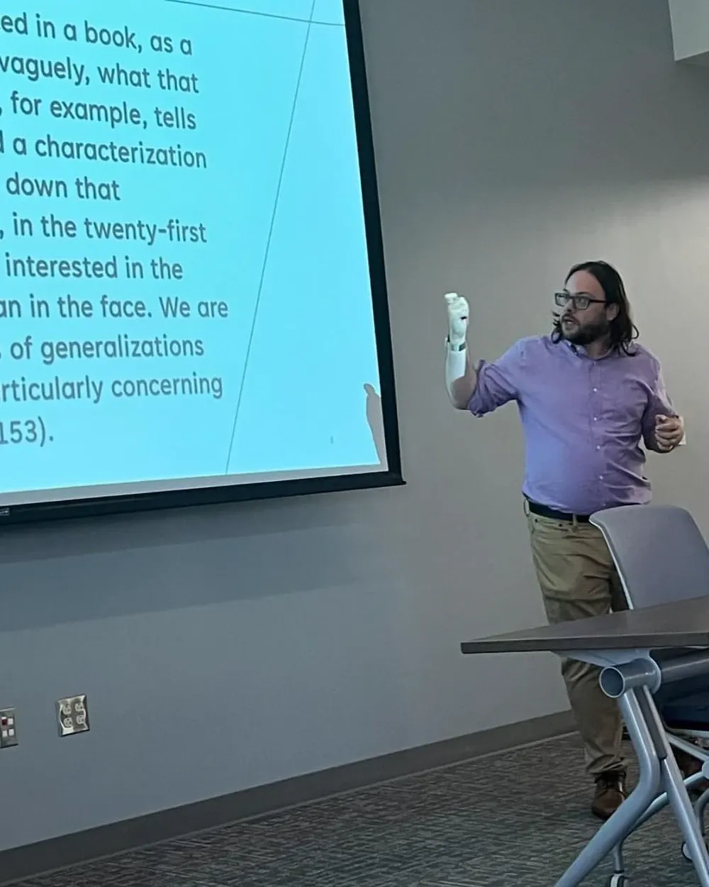 Eric Shay Howard gesturing mid-sentence in a lavender shirt and glasses, beside a projected slide quoting Charles Baxter on characterization.
